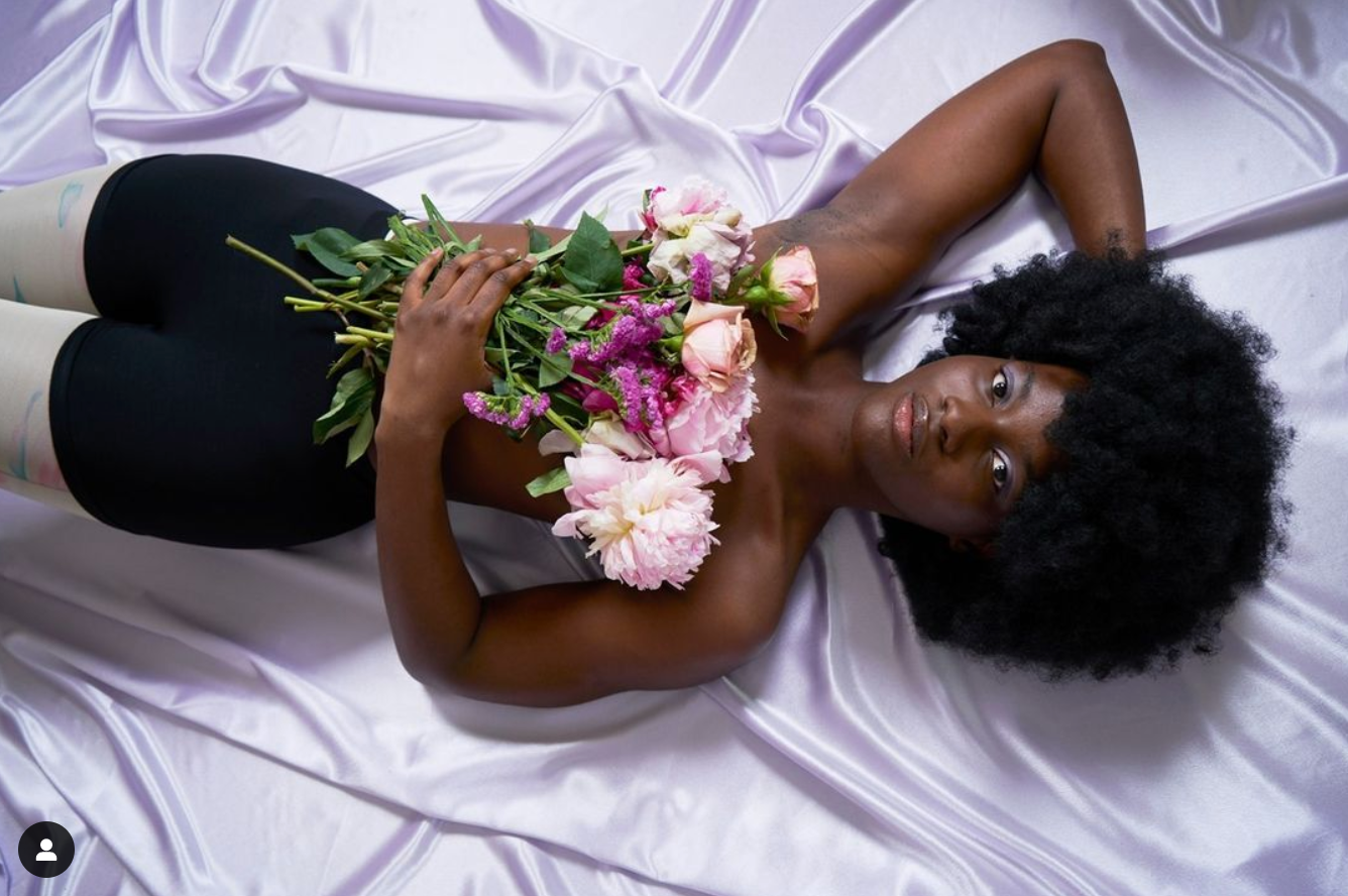 jeune femme à la peau noire en boxer allongée sur un drap en satin violet avec un main dans son afro et une autre portant un bouquet de fleur pour masquer sa poitrine nue